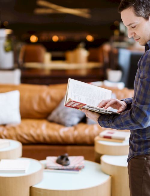 A person in a blue shirt is reading a book, standing in a modern living room with a brown leather sofa and round tables.