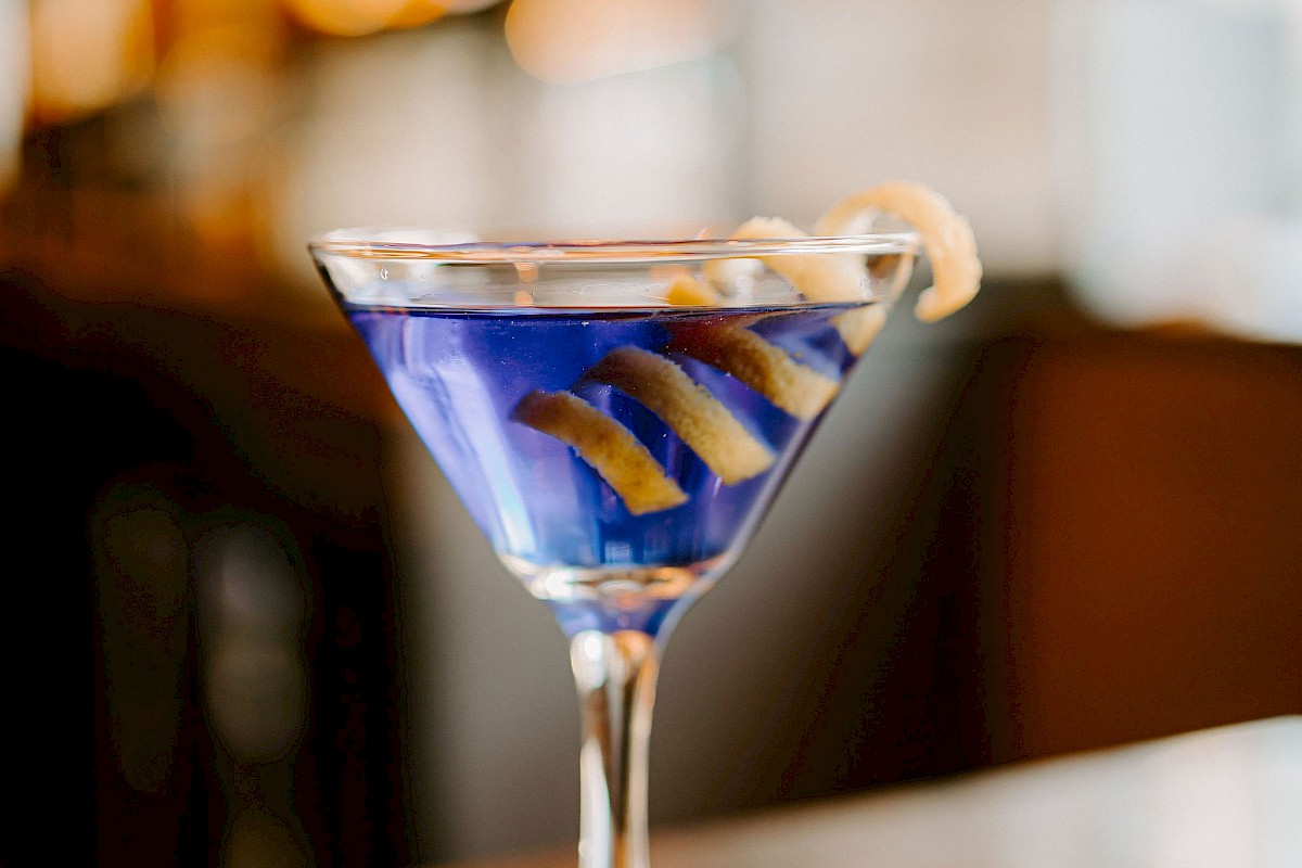 A cocktail in a martini glass with a blue liquid, garnished with a lemon twist, on a table. Warm blurred lights in the background.