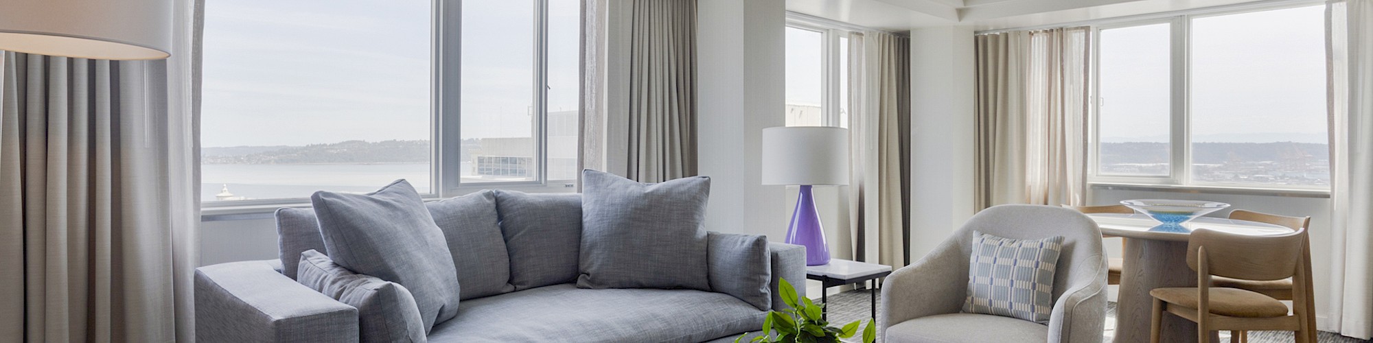 A modern, bright living room with large windows, grey sofa, beige chairs, a coffee table with a potted plant, and city views.