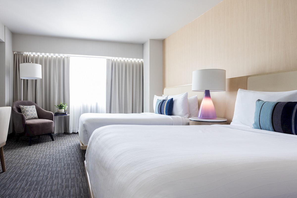 The image shows a bright, modern hotel room with two white beds, a round side table with a pastel-colored lamp, a purple armchair, a small table with a plant, and large window with curtains.