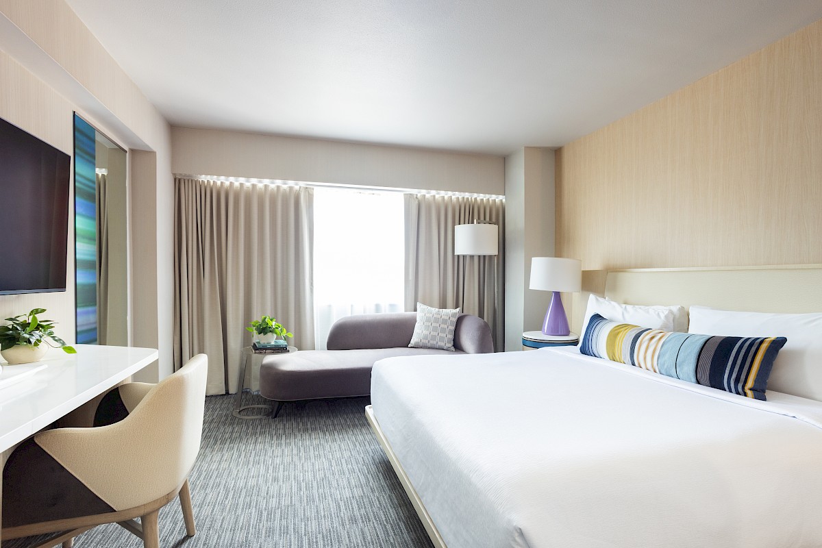 This is a bright, modern hotel room with a large bed, a sofa near the window, a desk, and decorative elements.