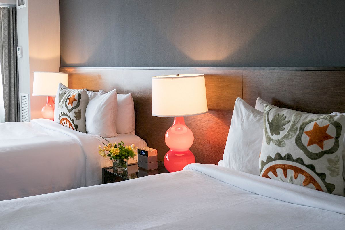 This image shows a modern hotel room with two neatly made beds, colorful pillows, bedside lamps, and a small flower vase.