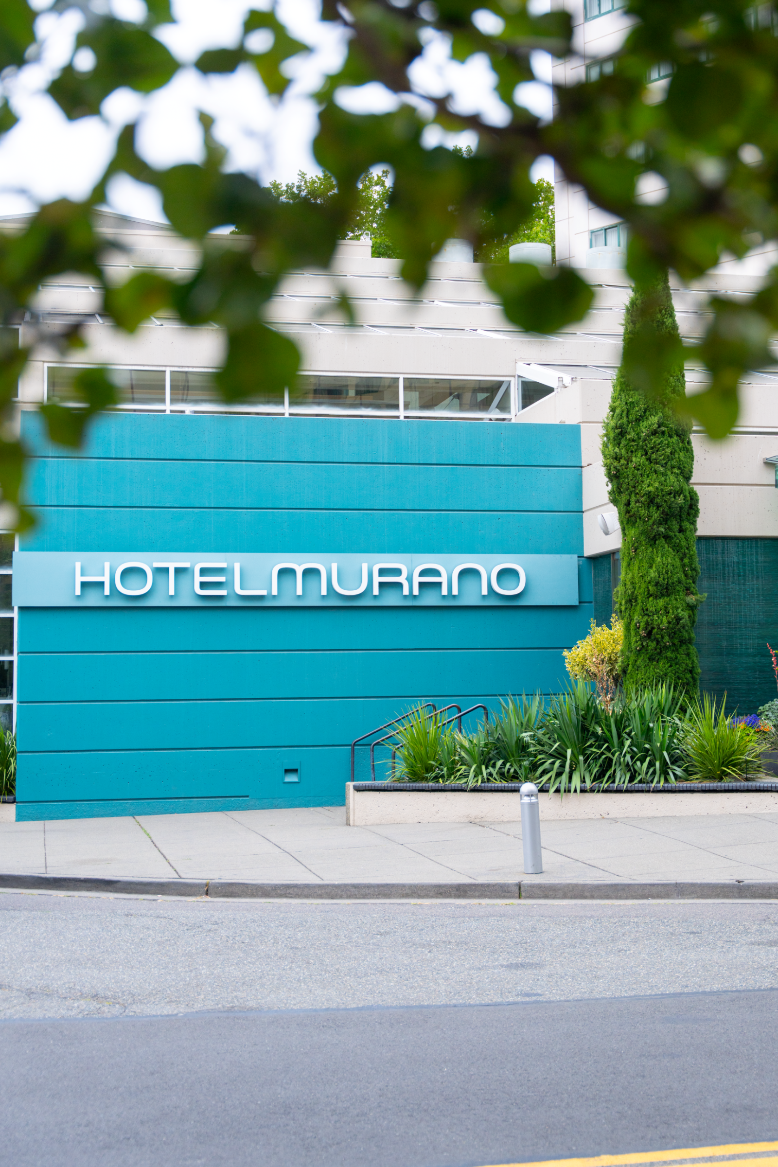 The image shows the exterior of Hotel Murano, partially obscured by green trees and plants.