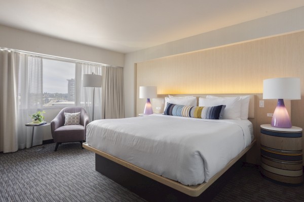 A cozy hotel bedroom with large bed, bedside lamps, armchair, plant, and city view from window.