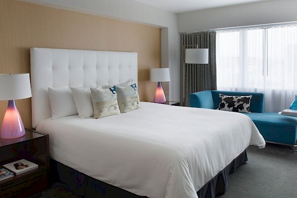 A modern bedroom has a king-sized bed with white linens, blue pillows, two nightstands, lamps, a turquoise chaise lounge, and natural light from a window.