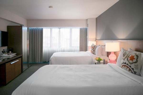 A modern hotel room with two double beds, a bedside lamp, a TV on a dresser, a window with curtains, and a coffee maker on the counter.