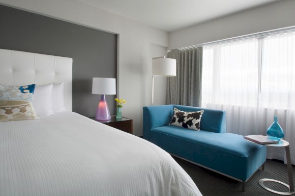 A modern hotel room with a bed, nightstand, lamp, blue chaise lounge, and a side table near a window with curtains ending the sentence.