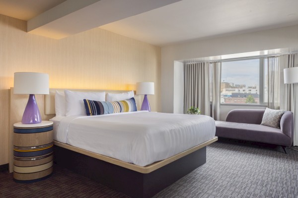 This is a modern hotel room with a large bed, side tables, purple lamps, a purple chaise lounge, and a city view through the window.