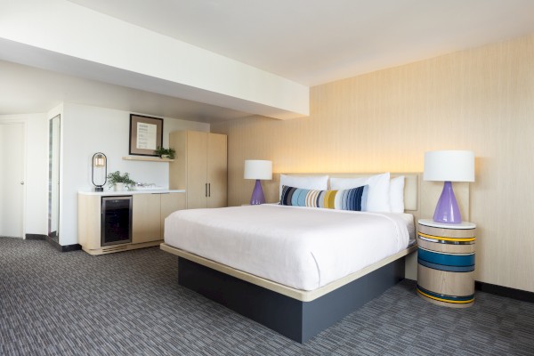 A modern hotel room with a large bed, colorful pillows, lamps, a mini fridge, and minimalist decor.