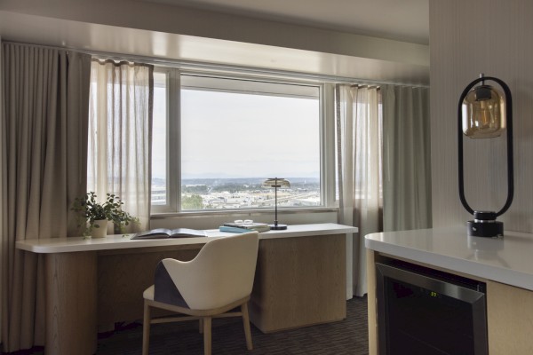 A bright hotel room with a desk by the window, natural light, a plant, books, a lamp, and a view of the city skyline in the background.