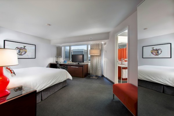 A modern hotel room with a large bed, a TV, a desk, a red lamp on a nightstand, a red chair, and an open bathroom door in the background.