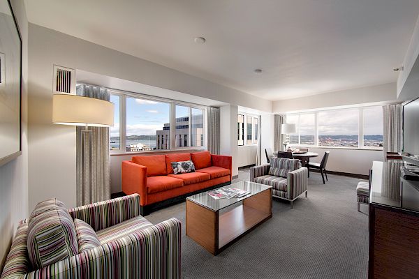 This image shows a bright, modern living room with a red sofa, striped chairs, a coffee table, and large windows offering city views, ending the sentence.