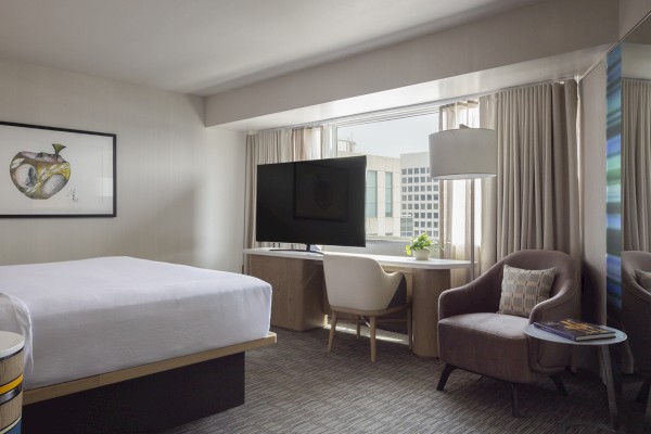 A modern hotel room features a bed, a TV, a desk, an armchair, and city views through large windows.