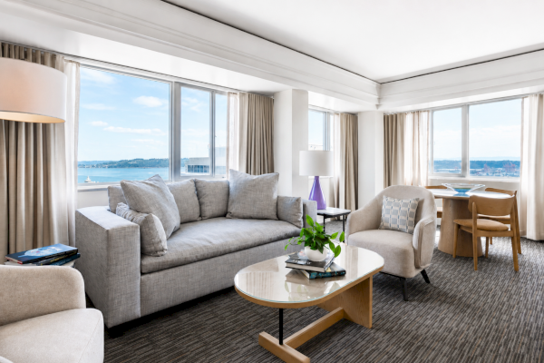 A bright, modern living room with large windows overlooking water, furnished with a gray sofa, armchair, coffee table, and decorative items.