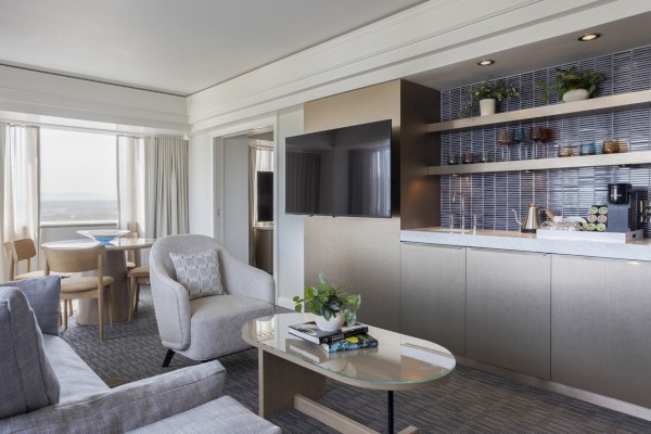A modern hotel suite features a sitting area, a kitchenette, a wall-mounted TV, and a large window with city views.