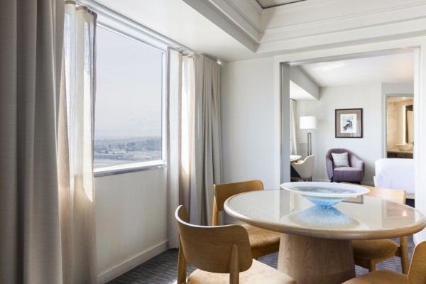 This image shows a dining area with a round table, chairs, curtains, and a view from a large window in a modern, well-lit hotel room.