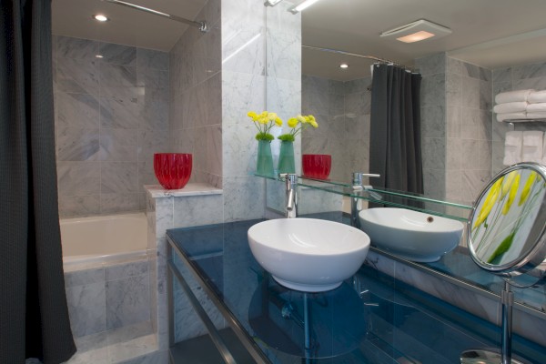 A modern bathroom with a glass-top sink, a round mirror, flowers in vases, and a bathtub with a dark curtain is visible.