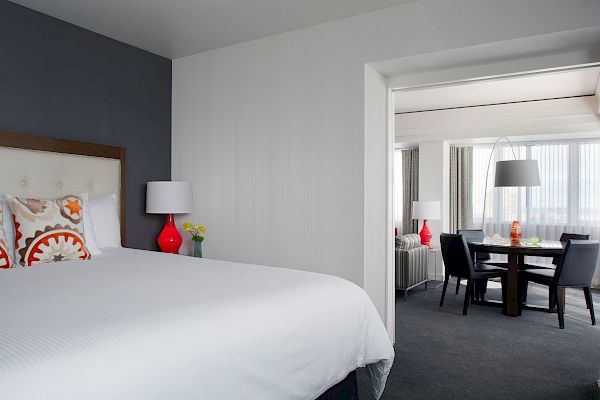 A modern and clean hotel room with a bed, pillows, and bedside lamps. A dining area with chairs and a table is visible through the doorway.