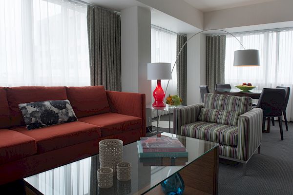 A modern living room with a red sofa, striped armchair, glass coffee table, floor lamp, and a dining table with chairs near the window.