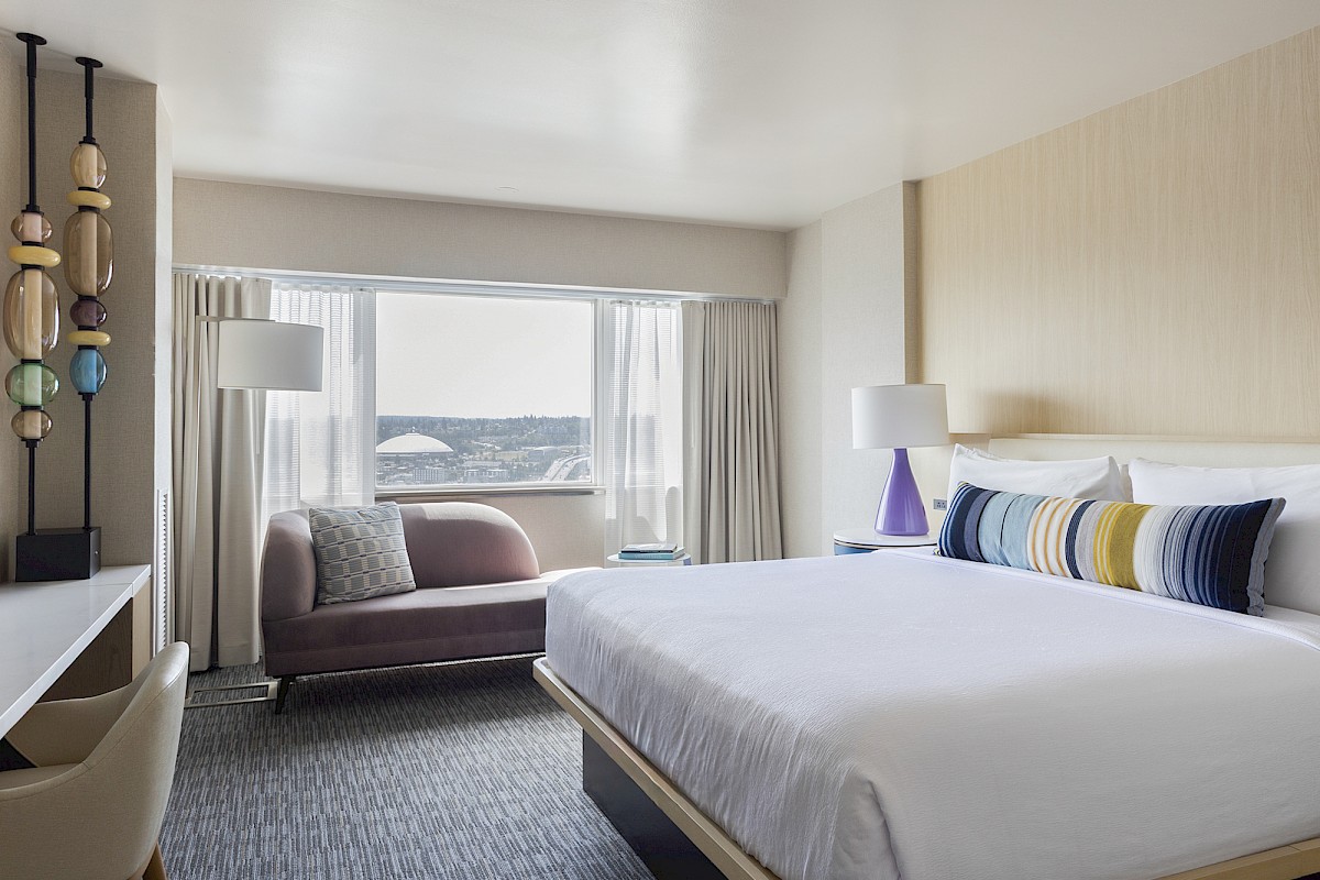 A stylish, modern hotel room with a large window, bed, colorful pillows, a lounge chair, and tasteful decor.