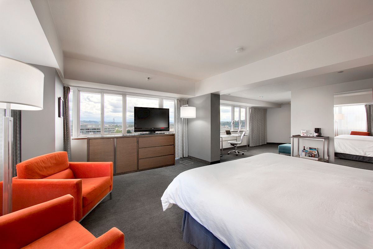 The image shows a modern hotel room with a bed, a flat-screen TV on a dresser, orange chairs, a desk and chairs in the back, and large windows.