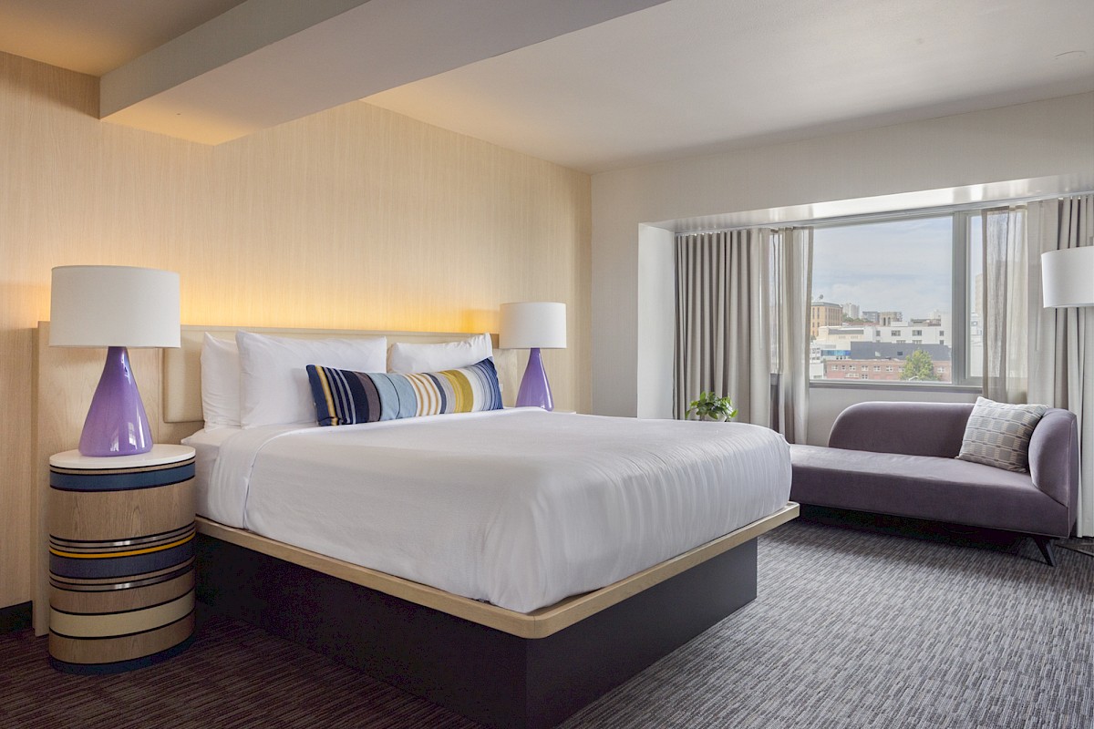 A modern hotel room featuring a large bed, purple lamps, a sofa by the window, and city views in the background.