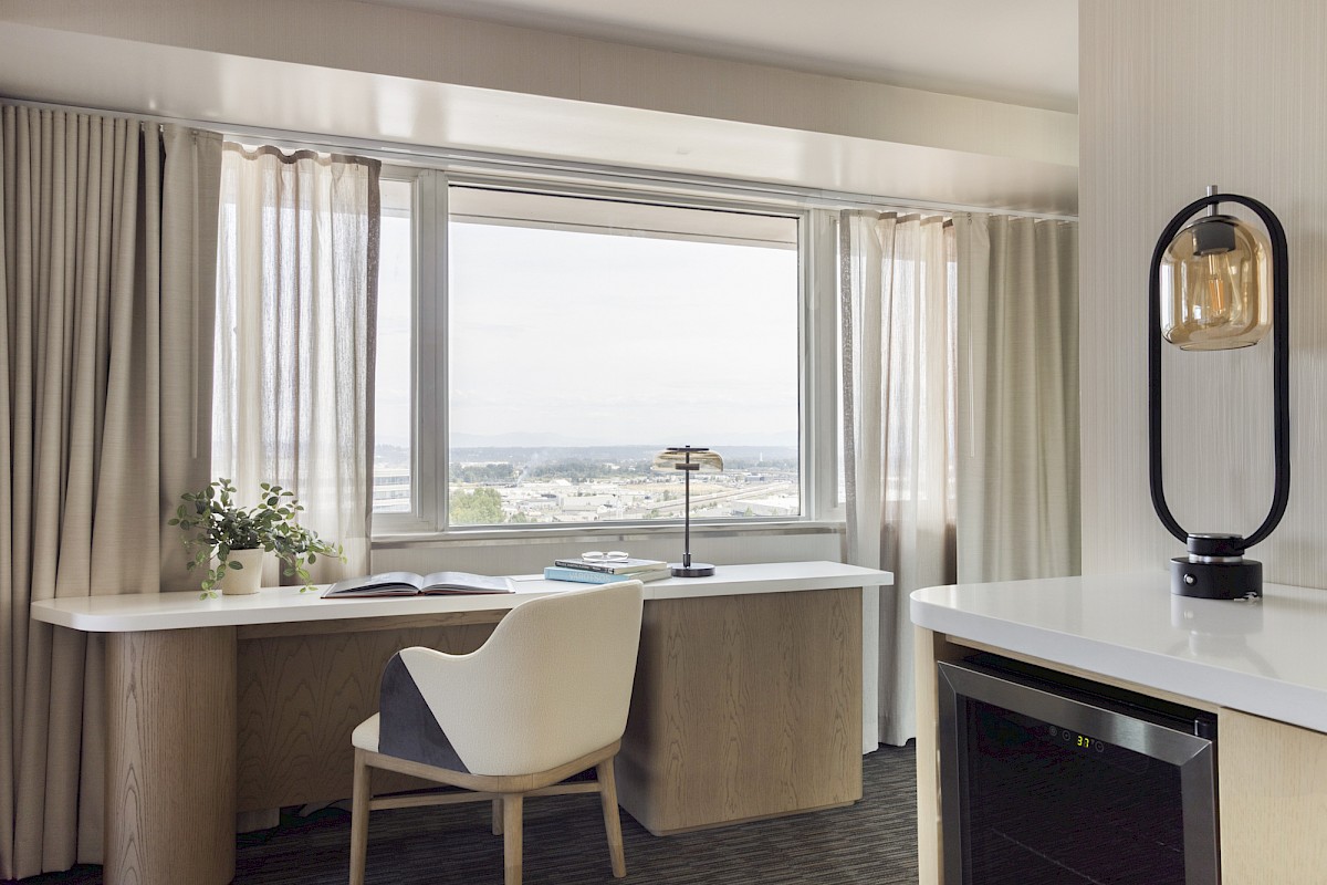 A cozy, modern hotel room with a large window, desk, chair, lamp, potted plant, and a mini fridge, offering a cityscape view.