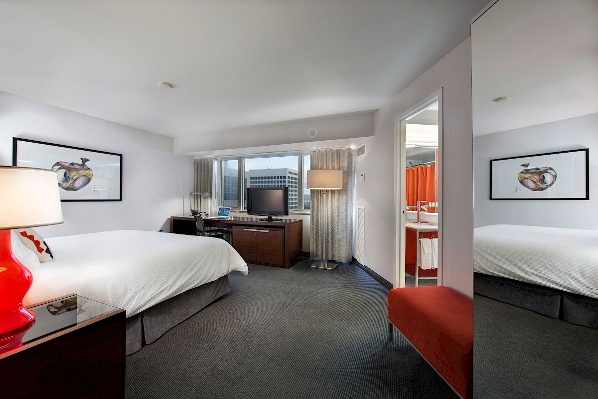 A modern hotel room with a double bed, flat-screen TV, desk, chair, red bench, and framed artwork, featuring a bathroom with an orange shower curtain.