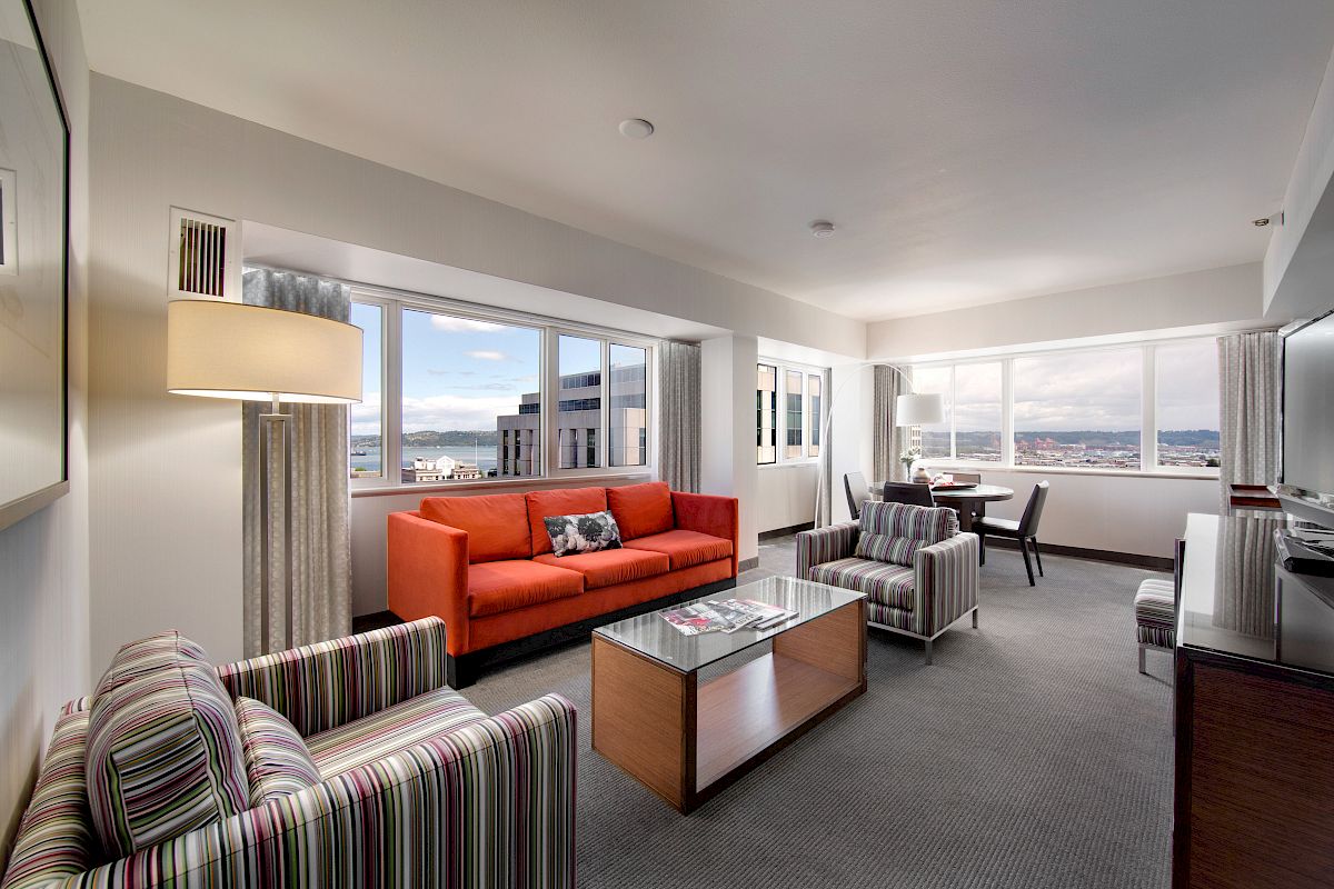 A modern hotel room features a red sofa, striped chairs, a glass coffee table, and large windows with city views, complemented by neutral tones.