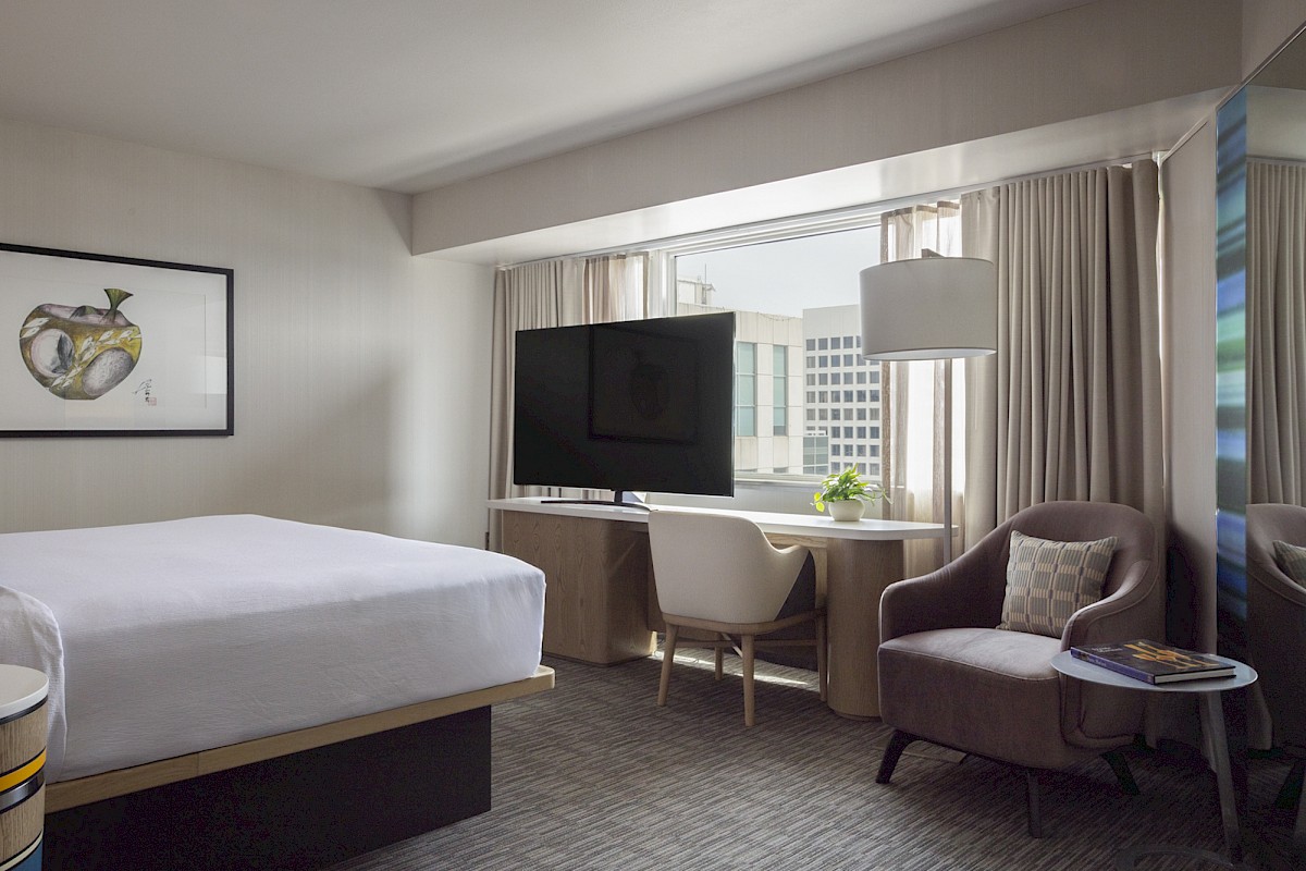 A modern hotel room with a bed, TV, desk, comfortable chair, and large window showing city buildings.