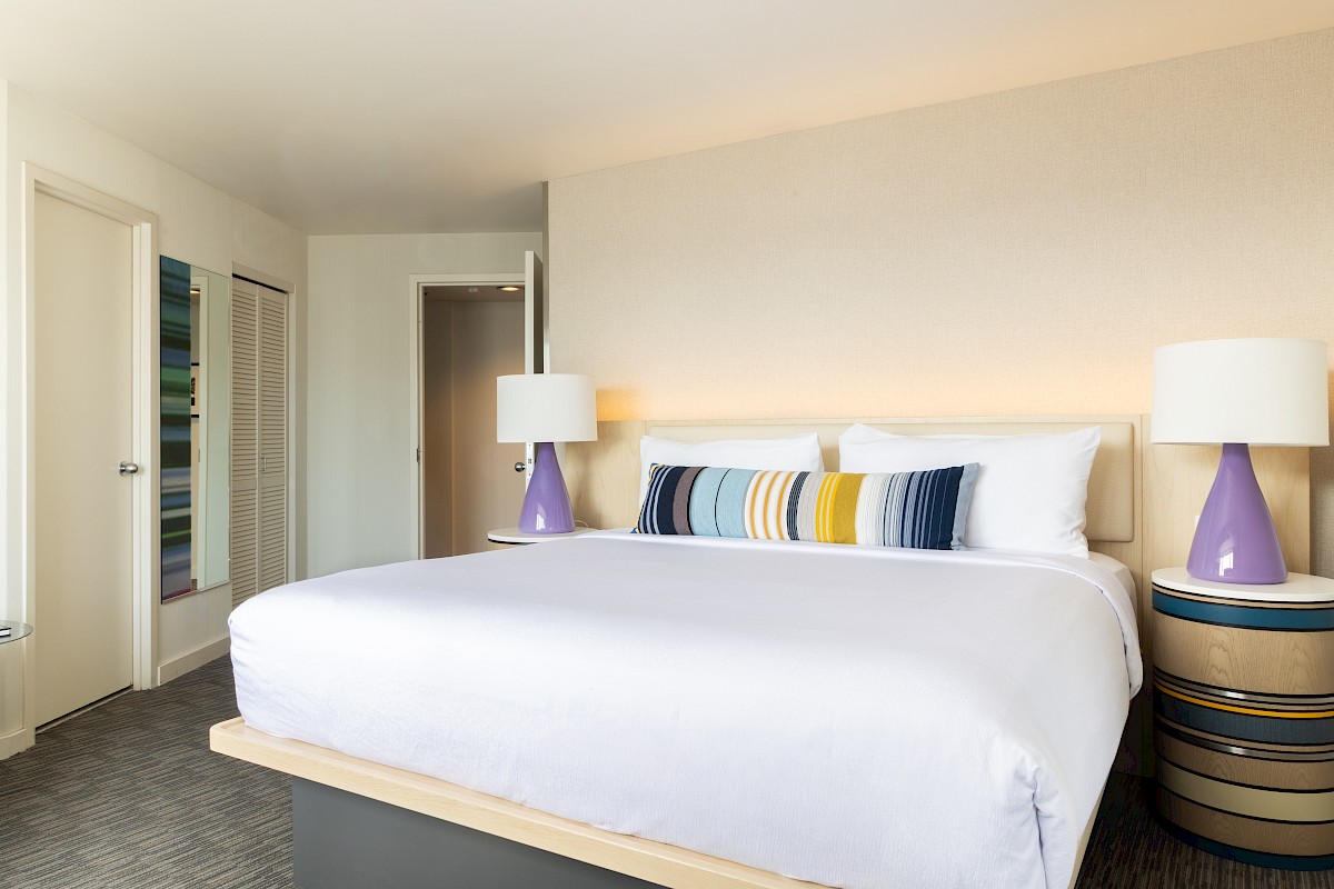 A hotel room with a large bed, colorful pillows, and matching lamps on side tables, featuring a simple, modern decor style.