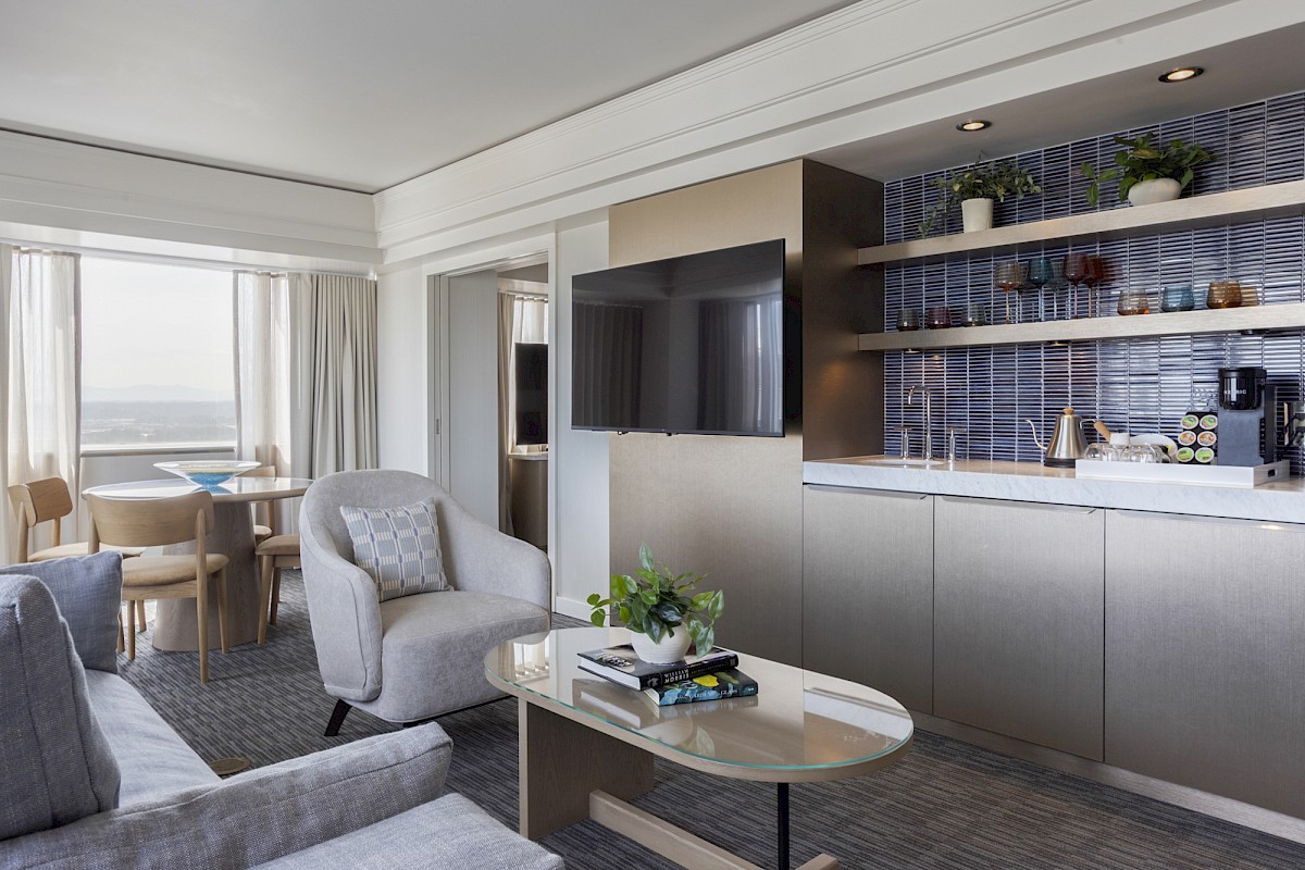 A modern, sunlit living space with a cozy seating area, a TV, a kitchen nook with shelves, and a view outside.