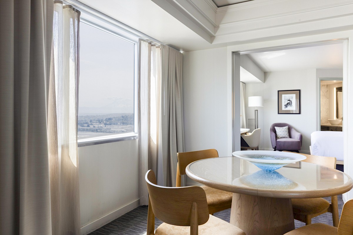 A modern hotel suite features a dining area with a round wood table and chairs, a large window with curtains, and a cozy sitting area with artwork.
