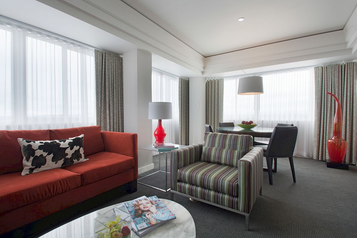 A modern living room with a red sofa, striped armchair, glass coffee table with magazines, and a dining area in the background.