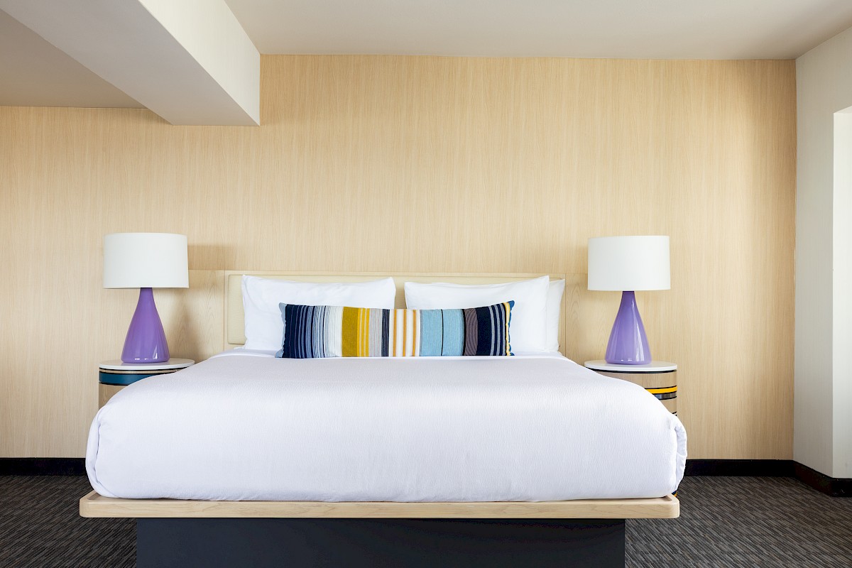 A neatly made bed with a decorative pillow, flanked by two purple lamps on side tables, against a light wood-paneled wall.