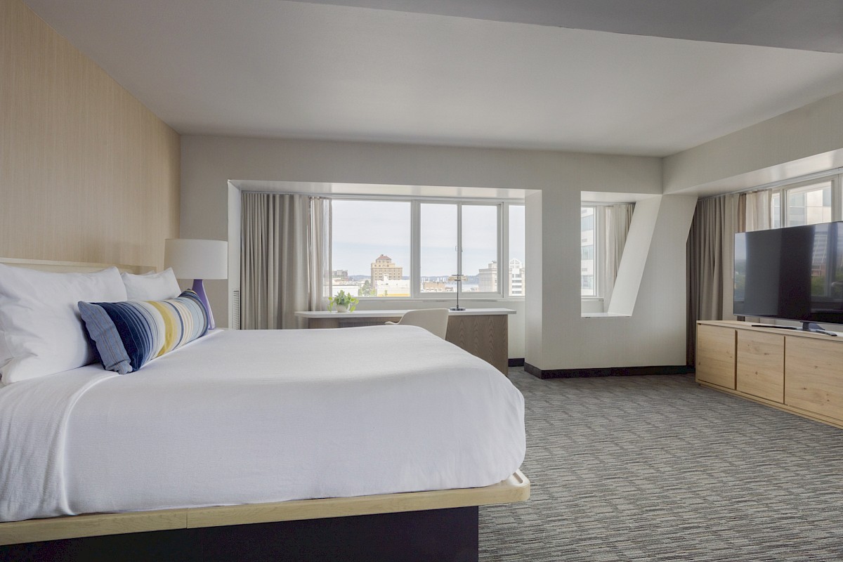 A modern, bright hotel room with a large bed, TV, desk, and a city view through big windows.