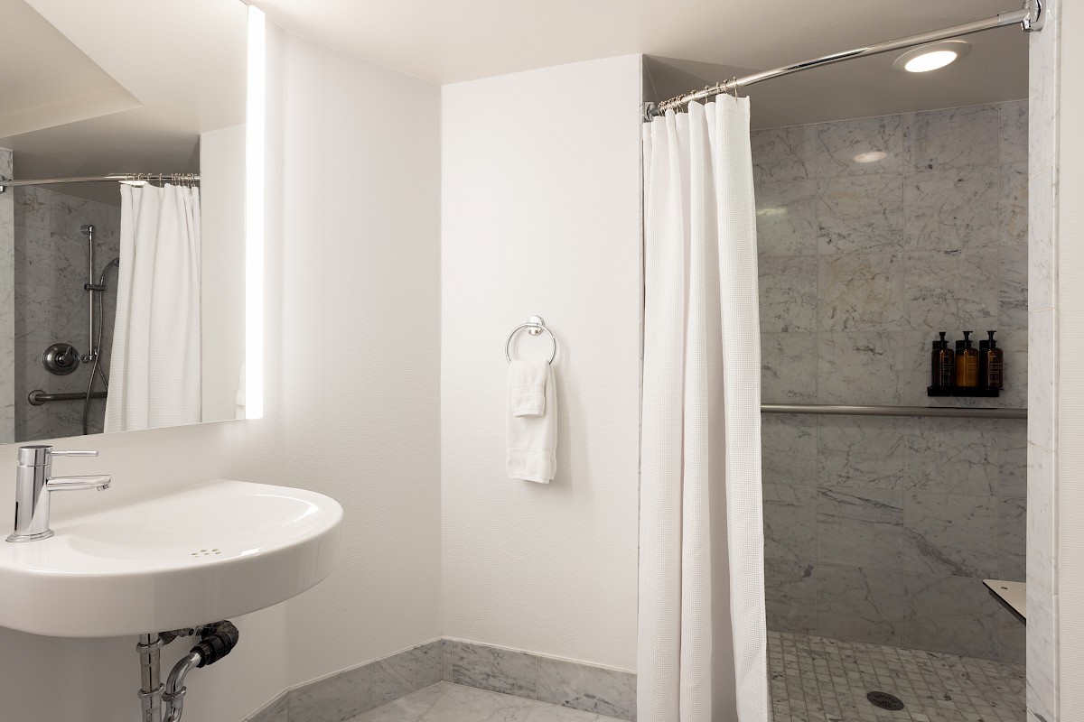 A minimalist bathroom featuring a sink with a mirror, a towel ring, and a marble shower with bottles on a shelf.