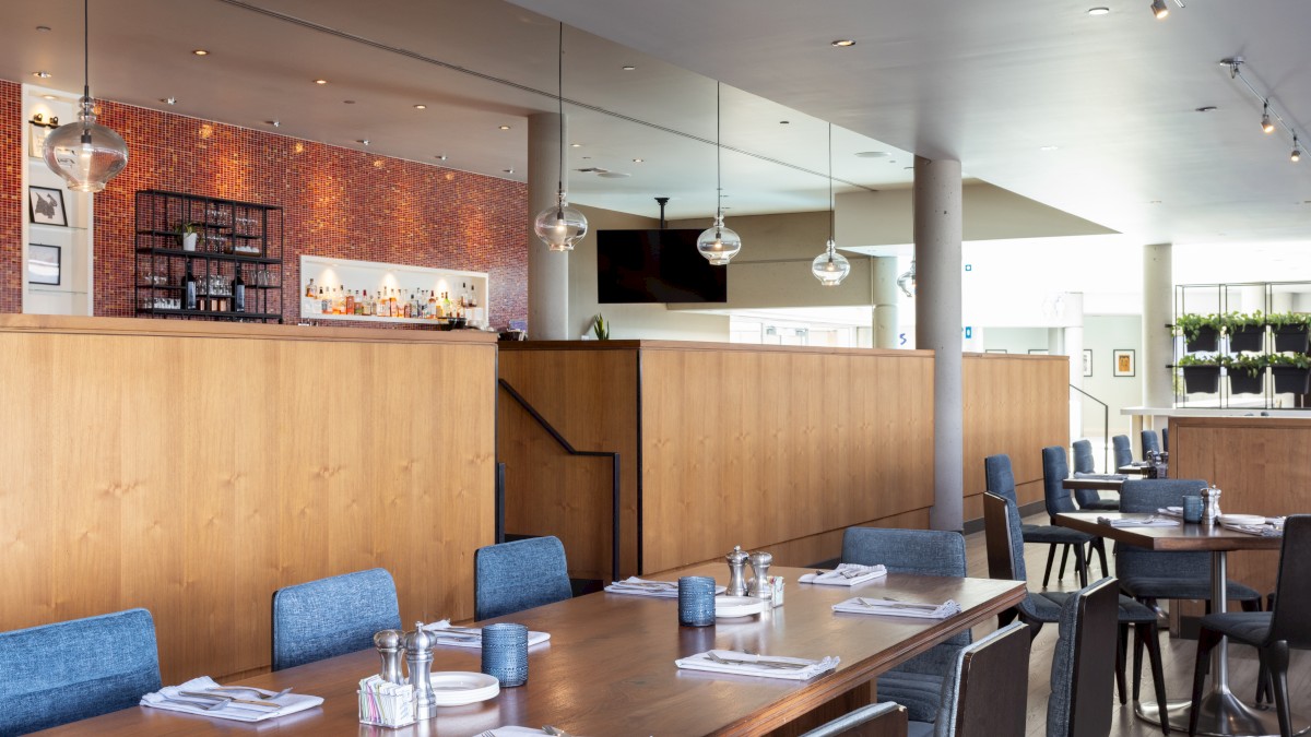 The image shows a modern, upscale restaurant interior with wooden tables, blue chairs, a bar area with bottles, and decorative lighting.