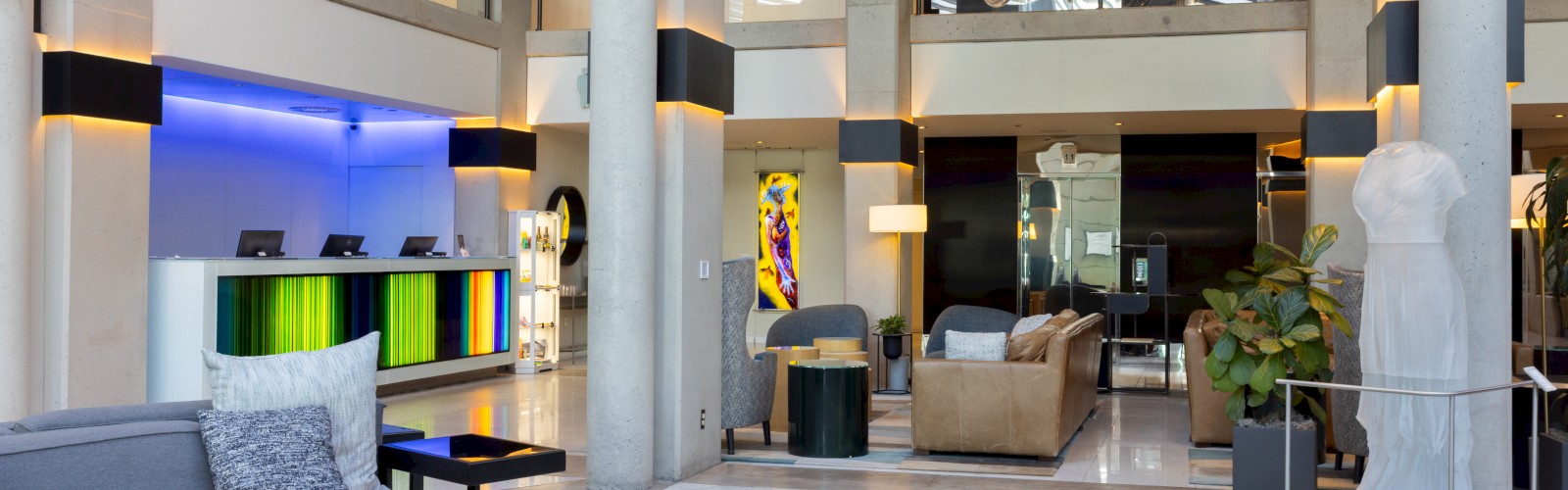 This image shows a spacious, modern hotel lobby with stylish furniture, sculptures, plants, a reception desk, and decorative lighting.