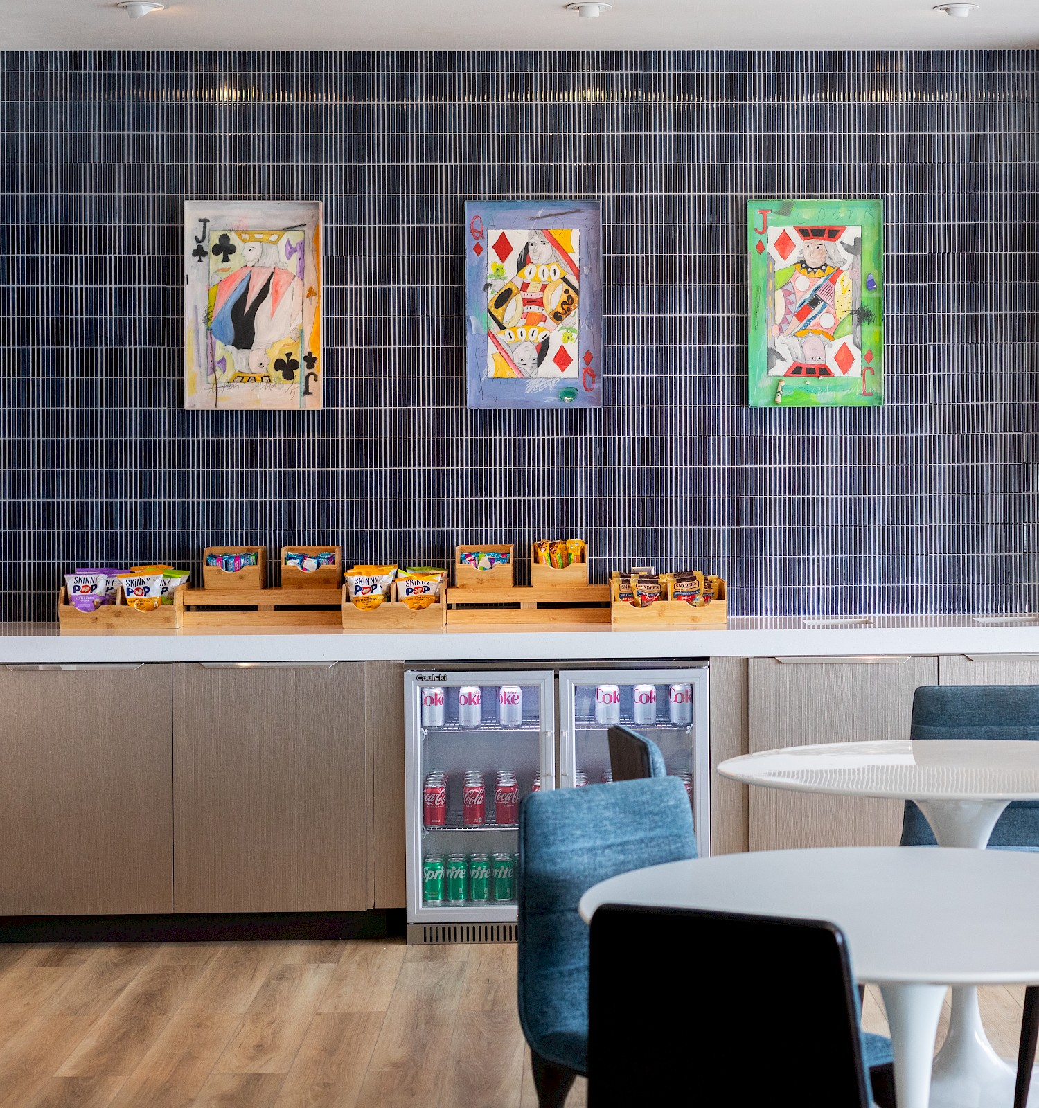 A modern, cozy dining area with playing card artwork on the wall and snacks on the counter, with a small fridge stocked with sodas.