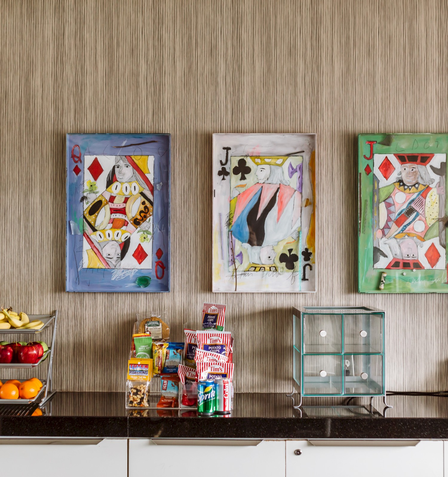 The image shows a countertop with fruits and snacks, and three colorful framed playing card artworks on the wall in the background.