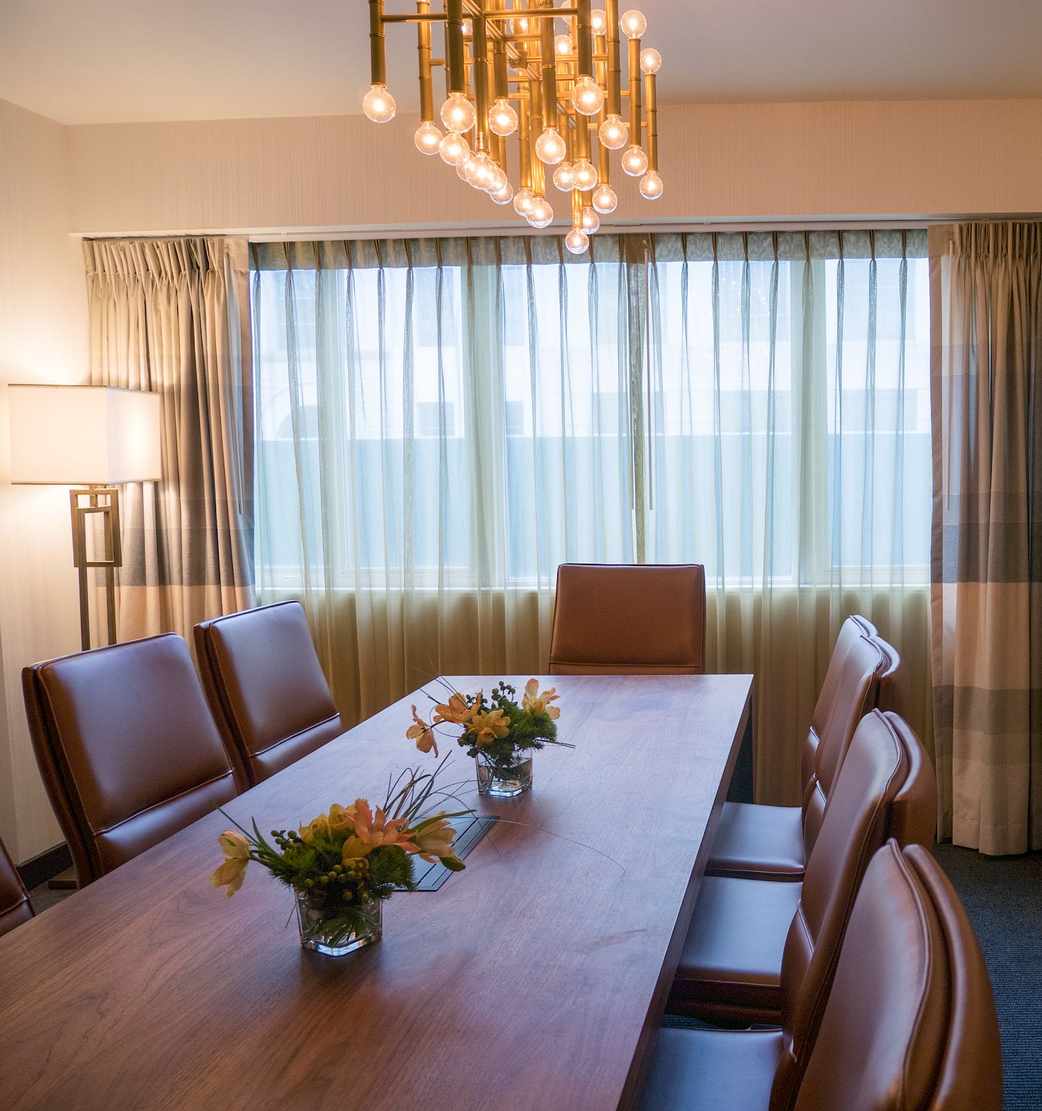 A room with a wooden table, eight chairs, chandelier, window with curtains, cabinet, lamp, flowers in vases, and framed artwork on the wall.