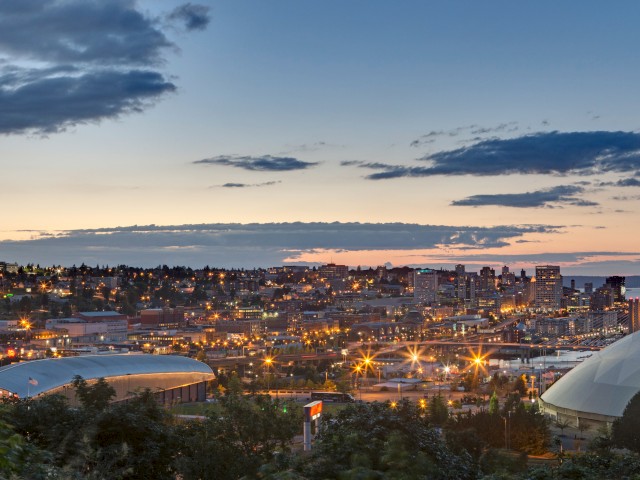 A cityscape at dusk with illuminated buildings and two large dome structures in the foreground under a blue and cloudy sky.