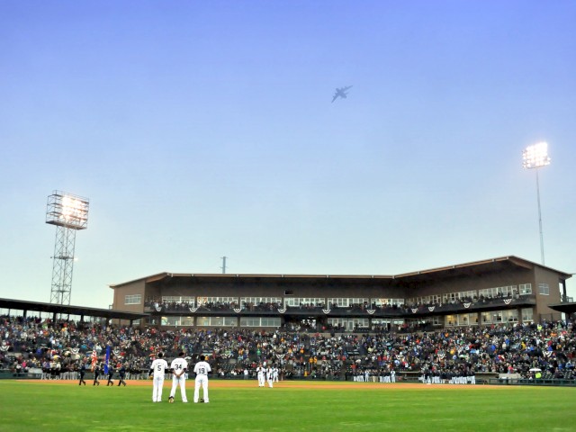A baseball game is underway with players on the field, a packed stadium, and a plane flying overhead in the clear sky.