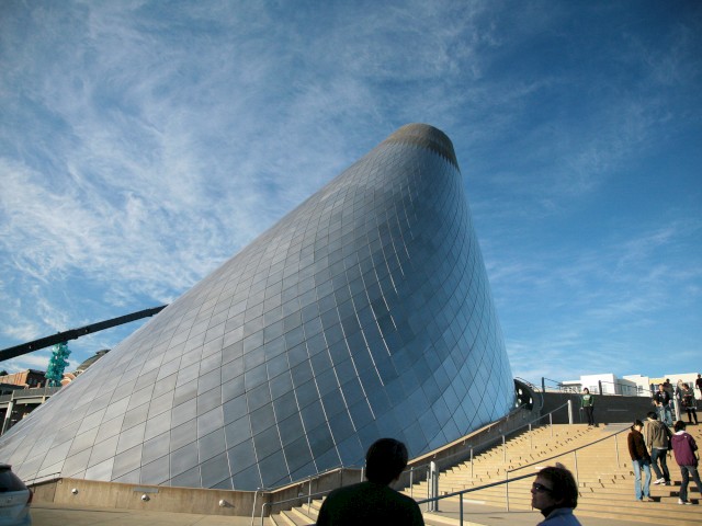 A modern, conical building with a metallic exterior under a blue sky, people are walking nearby on a staircase.