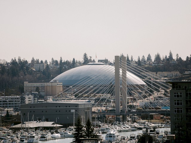 The image shows a waterfront with boats, a large dome structure, and a cable-stayed bridge with trees in the background, ending the sentence.