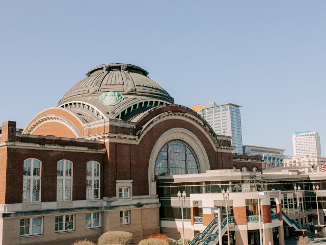 A large, historic building with an arched roof and multiple windows, surrounded by modern skyscrapers under a clear blue sky.