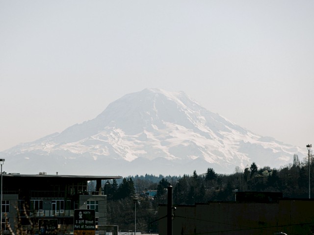 The image shows a distant, snow-capped mountain with buildings and trees in the foreground, under a clear sky.