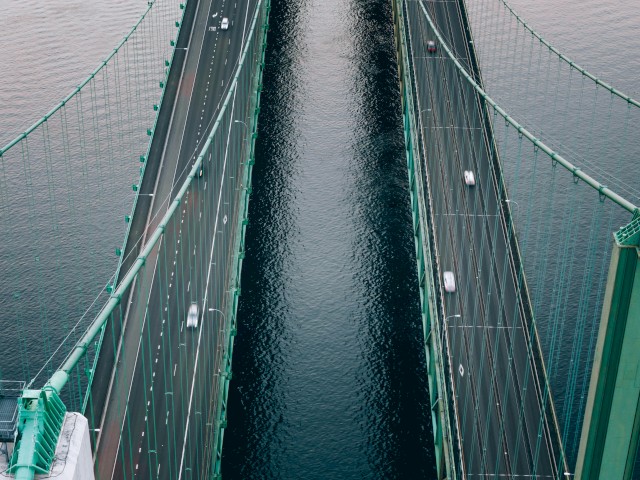 Aerial view of a suspension bridge with two parallel roadways and sparse traffic, crossing over water.