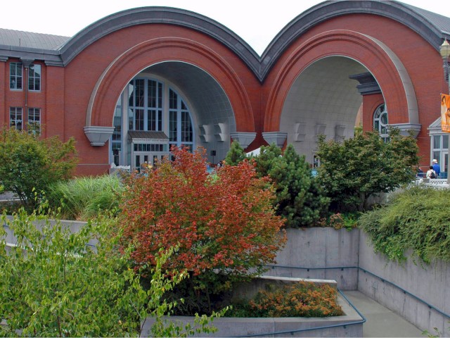 This image shows a large brick building with arched entrances and greenery in the foreground, likely part of a public or historical site.
